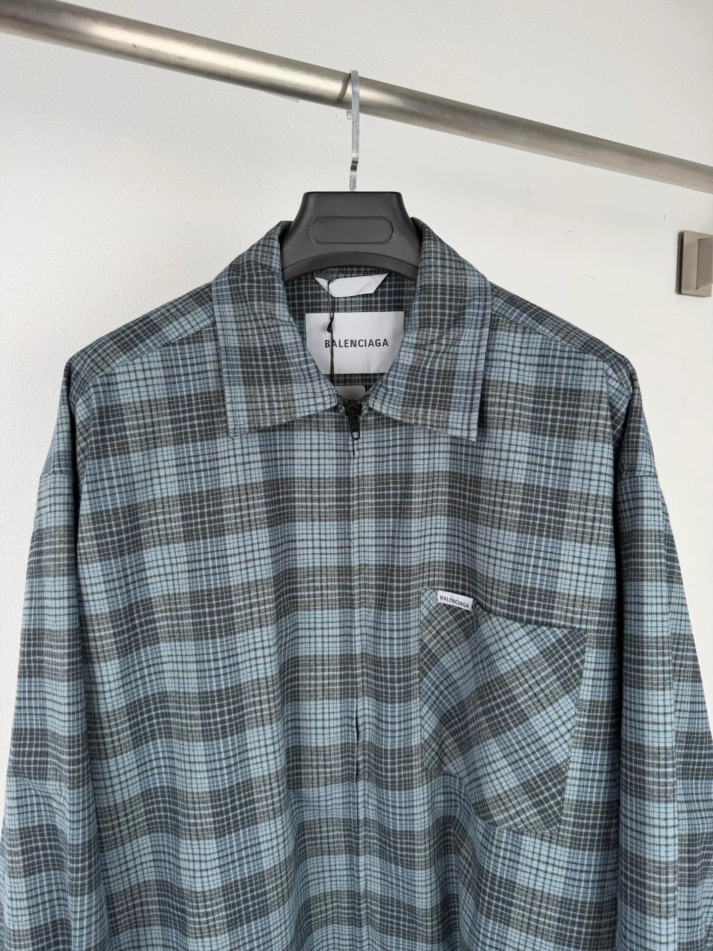 BALENCIAGA/balenciaga blue and gray plaid zipper pocket logo quilted jacket combines a three-layer structure of plaid woven fabric, filling cotton, and lining, and integrates plaid alignment technology and quilted garment production standards. There are multiple process difficulties in the whole process. The surface layer of the body is made of blue-gray Retro plaid fabric, and the cutting machine production strictly follows the global grid standards. The arrangement direction of the cutting pieces is fixed and cannot be changed at will. The horizontal and vertical lines of shoulder seams, side seams, and zipper placket joints must be consistent and coherent. The left chest patch pocket is cut independently, and the bag check pattern must be aligned with the front reference pattern. Slight deviations will destroy the visual integrity and directly increase fabric loss and cutting time. The most difficult part of the style is the three-layer composite sewing. The outer plaid fabric, filling cotton, and lining fabric have different physical properties, and the preshrinking processing parameters need to be adjusted separately. Pressure and temperature control are crucial during lamination operations. Once the parameters are out of balance, it can easily cause fabric stretch and distortion, plaid dislocation, and wrinkles and bulges in garments. At the same time, it is necessary to reasonably plan the quilting line to avoid the visual beauty of the grid pattern from being cut by the lines on the basis of firmly fixing the cotton layer and preventing cotton from running away. The placket adopts a centered zipper structure. It is difficult to sew the zipper tape and the quilted panels. It is required that the width of the left and right plackets be equal, the zipper routing is straight and smooth, and there is no lag in opening and closing. When the shirt lapel is filled with cotton, the thickness of the cotton needs to be controlled to prevent the collar from becoming bloated and unnaturally folded. The metal nameplate of the chest pocket is sewn at fixed points to avoid pulling the fabric during the operation and ensure that the plaid pattern is smooth and not deformed. The quilted material increases the overall thickness of the fabric. When sewing, you need to continuously adjust the presser foot pressure and stitch length to adapt to changes in sandwich thickness. The low-temperature and gentle ironing process is used for post-finishing to prevent the plaid from fading and the cotton layer from clumping due to high temperatures. From the precise alignment of cutting pieces, three-layer fabric pretreatment, composite quilting to garment finalization inspection, the processes are interlocking and the error tolerance rate is extremely low, testing the accuracy of plate making, equipment control and turning technology. Size:XS S M L